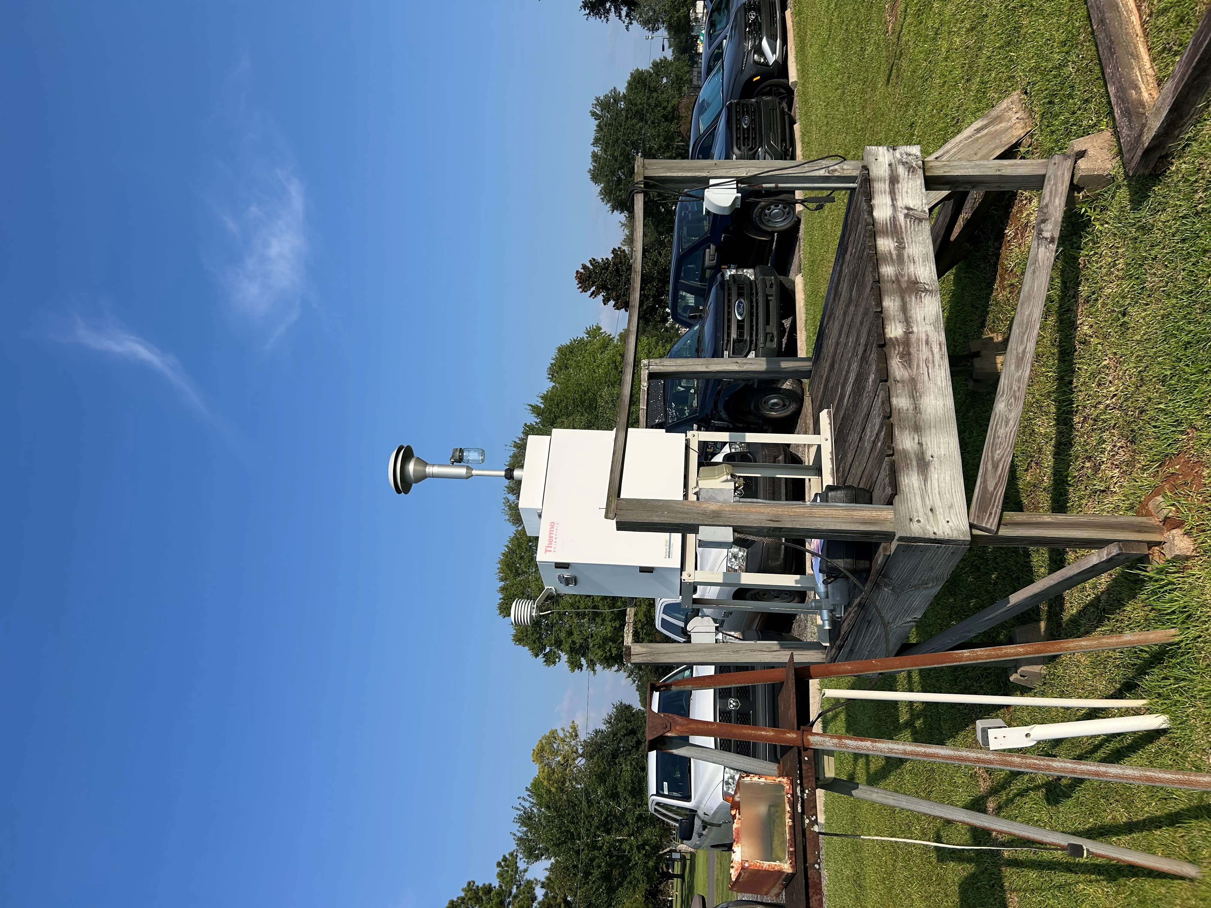 A wooden platform with a piece of air monitoring equipment on top and vehicles in the background.