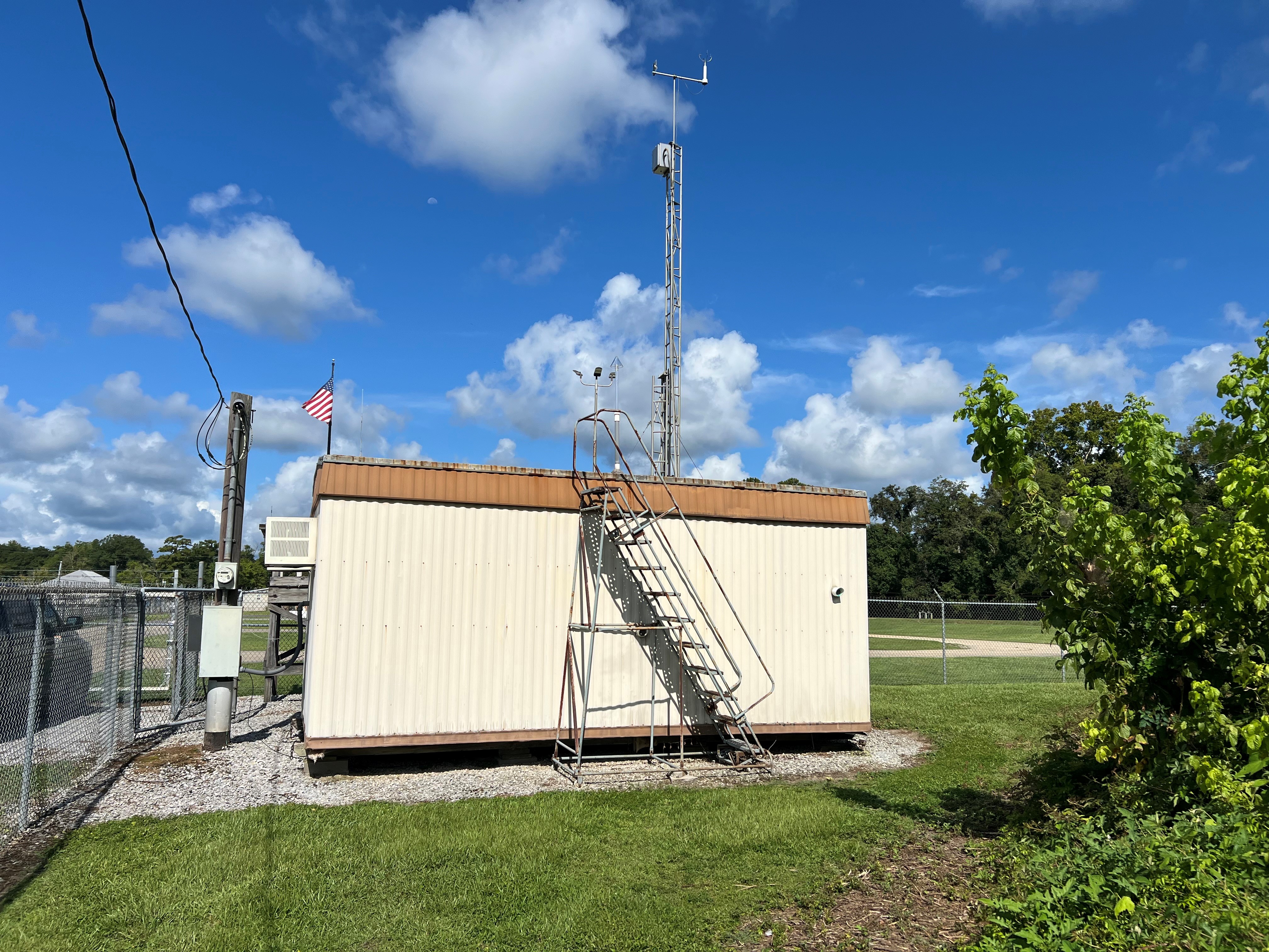 A tan metal building on a gravel pad surrounded by grass with a ladder to the roof and a meteorological tower in the back, a bush to the right and blue skies in the background.