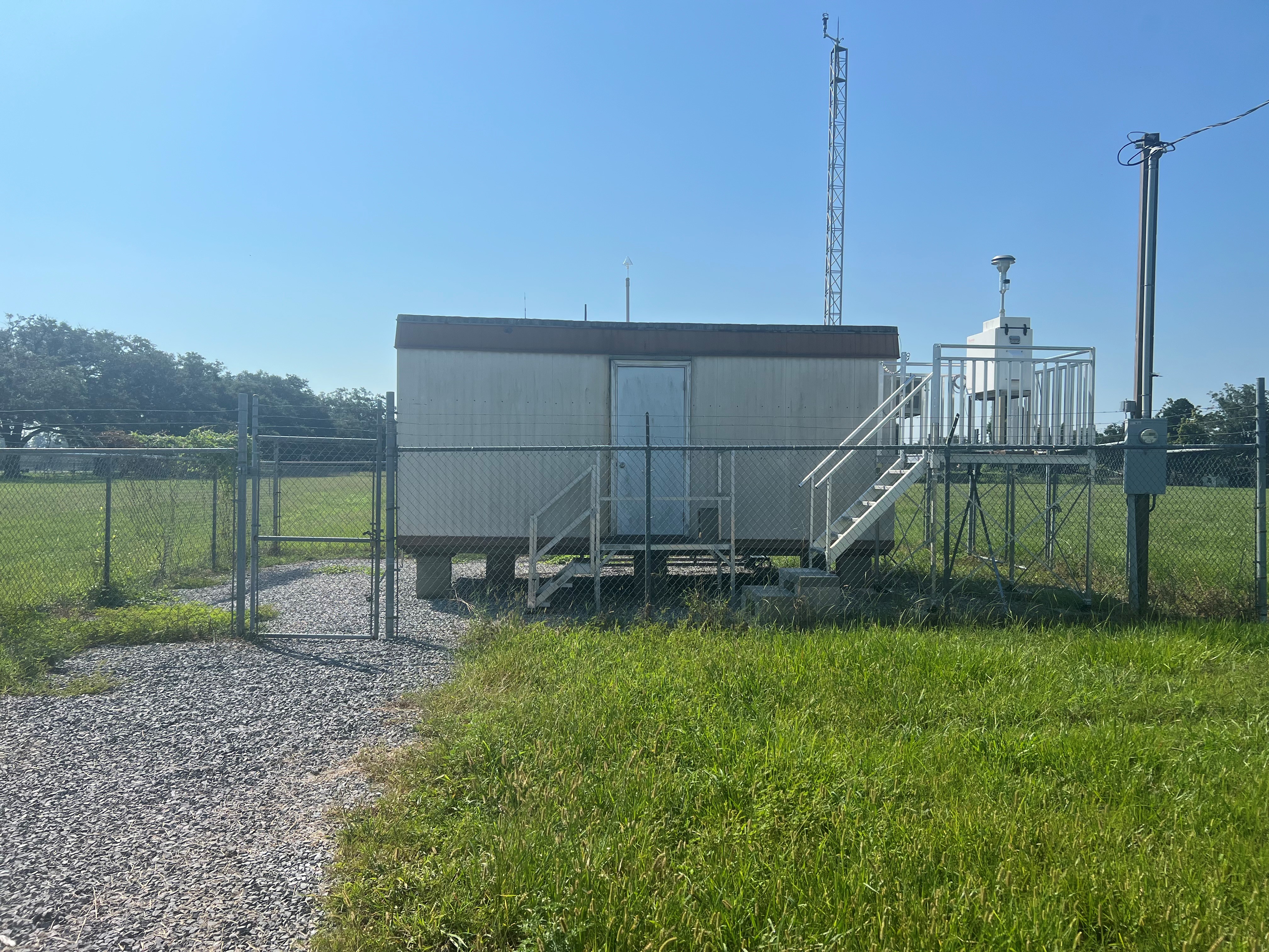 A metal building on top of gravel inside a fence, with a meteorological tower in the back right and a metal platform holding a piece of air monitoring equipment in front of the building.