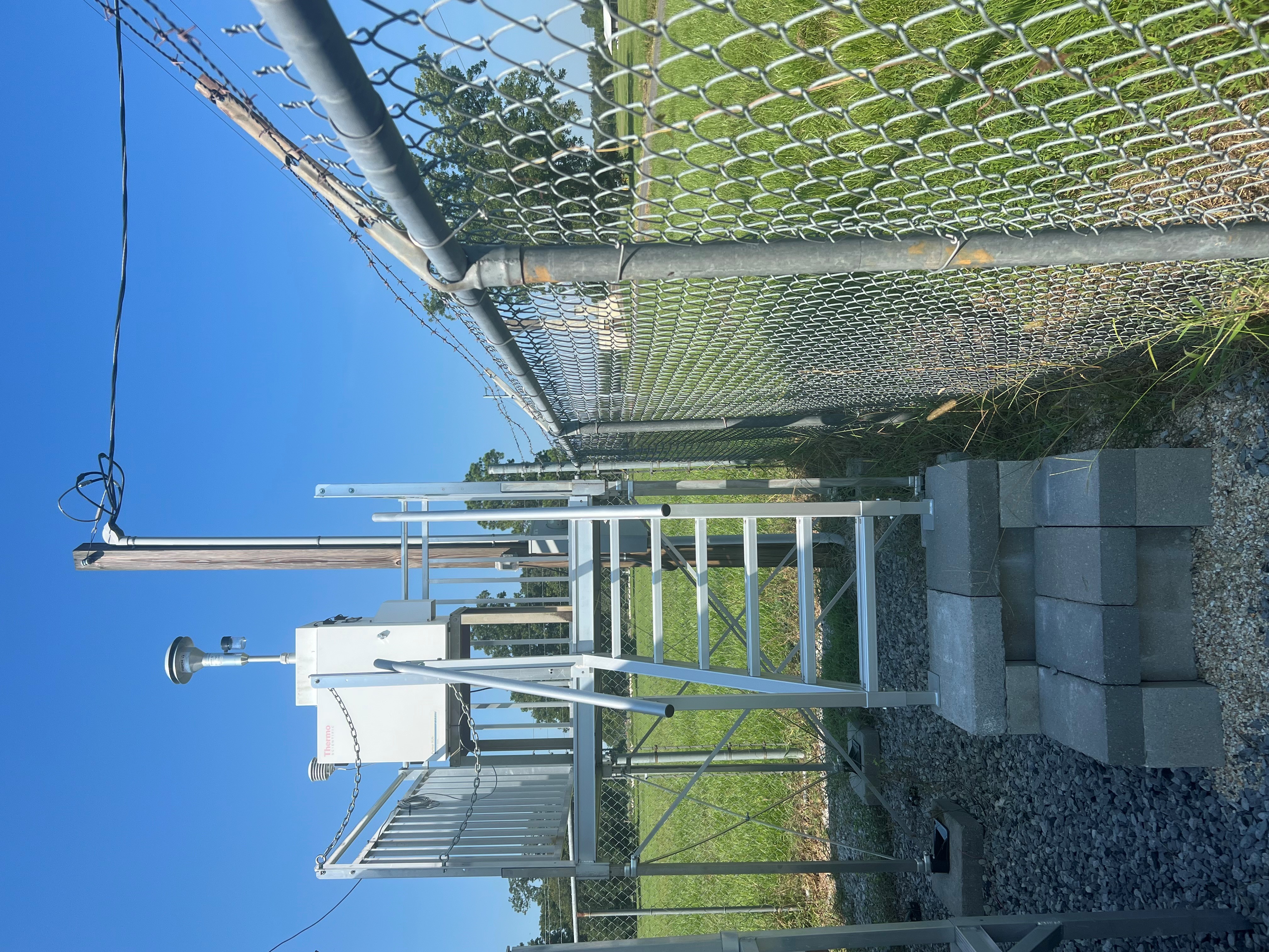 A metal staircase with a piece of air monitoring equipment on top, with a chain link fence to the right and in the back.