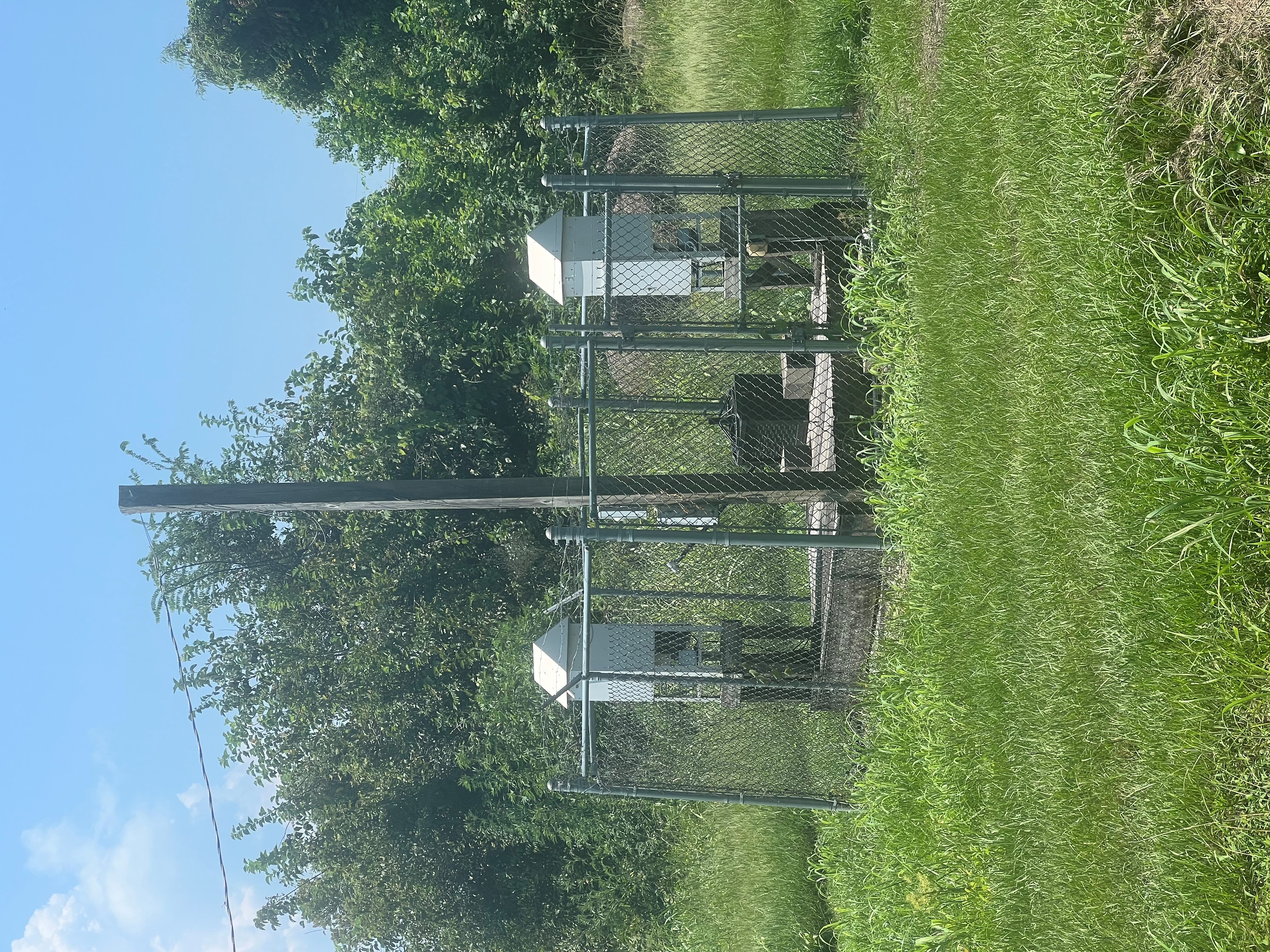 A grassy piece of land with a small area enclosed by a chain link fence with two metal air samplers inside.