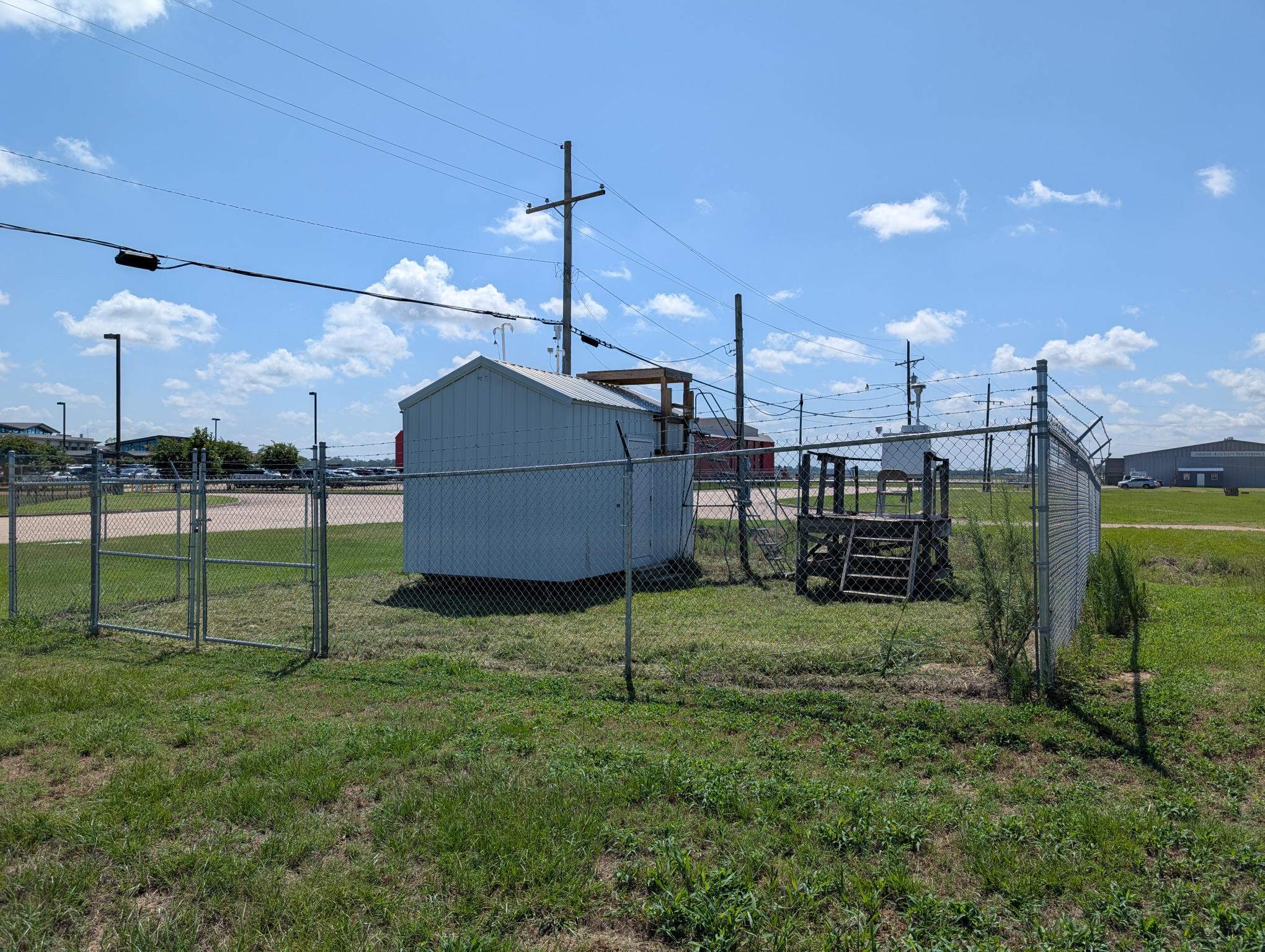 A gray metal building surrounded by a chain link fence, on a grassy piece of land with a wooden platform holding a piece of air monitoring equipment to the right of the building.