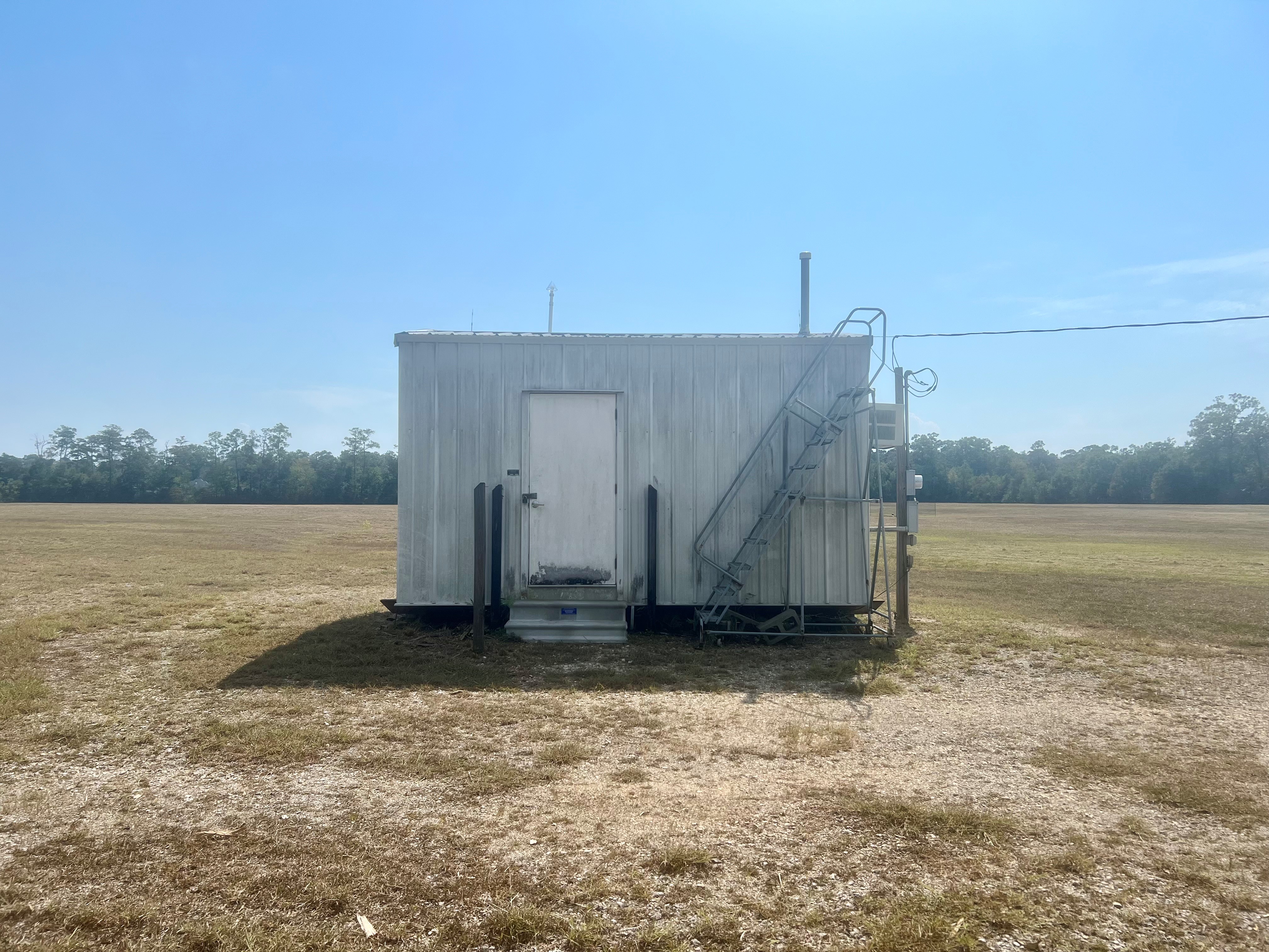 A small metal building with concrete steps and wooden rails on a bare piece of land with grass and trees in the background.
