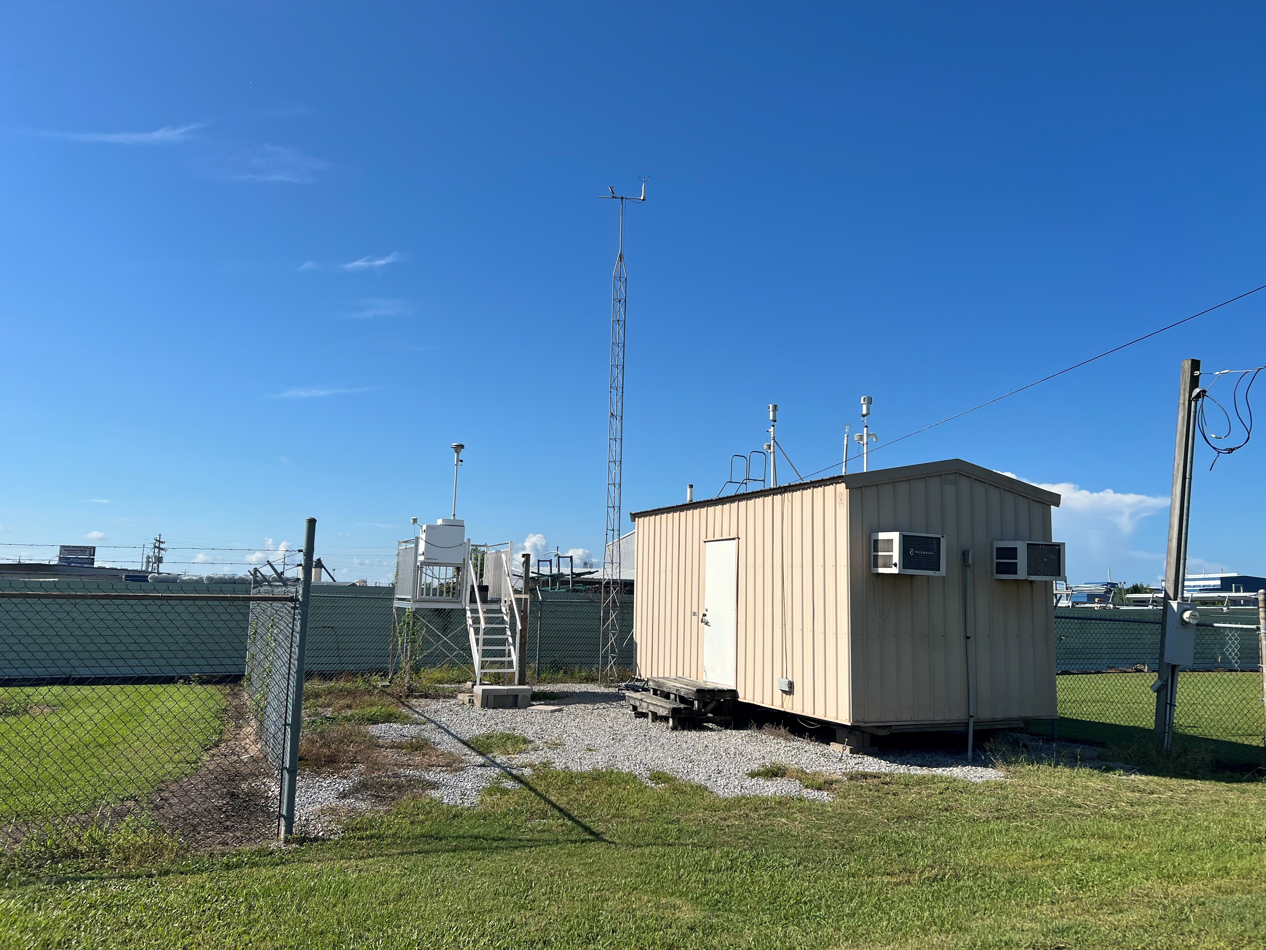 A tan metal building on a gravel pad with a chain link fence surrounding it on three sides and a metal platform with a piece of air monitoring equipment on top.