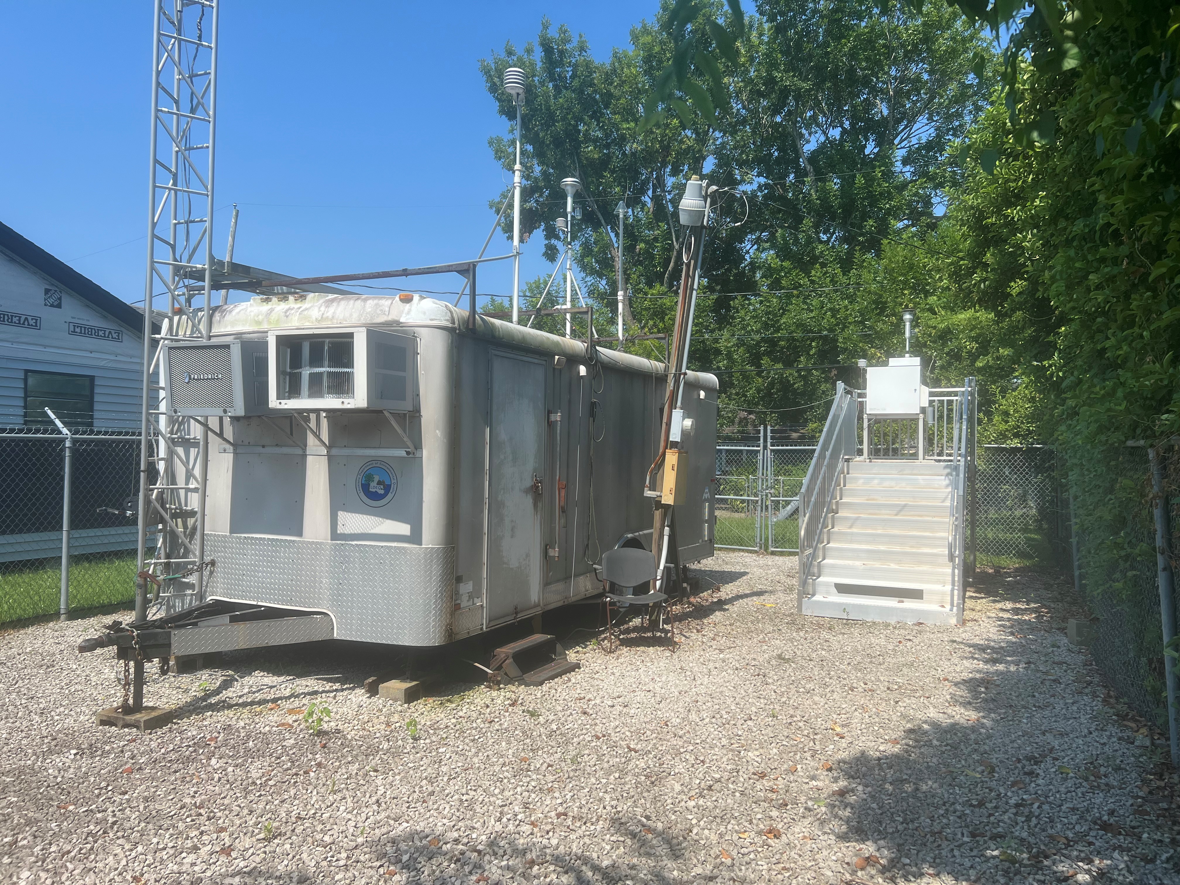 A white enclosed metal trailer inside a grassy, fenced in area, with a metal platform holding a piece of air monitoring equipment to the right.