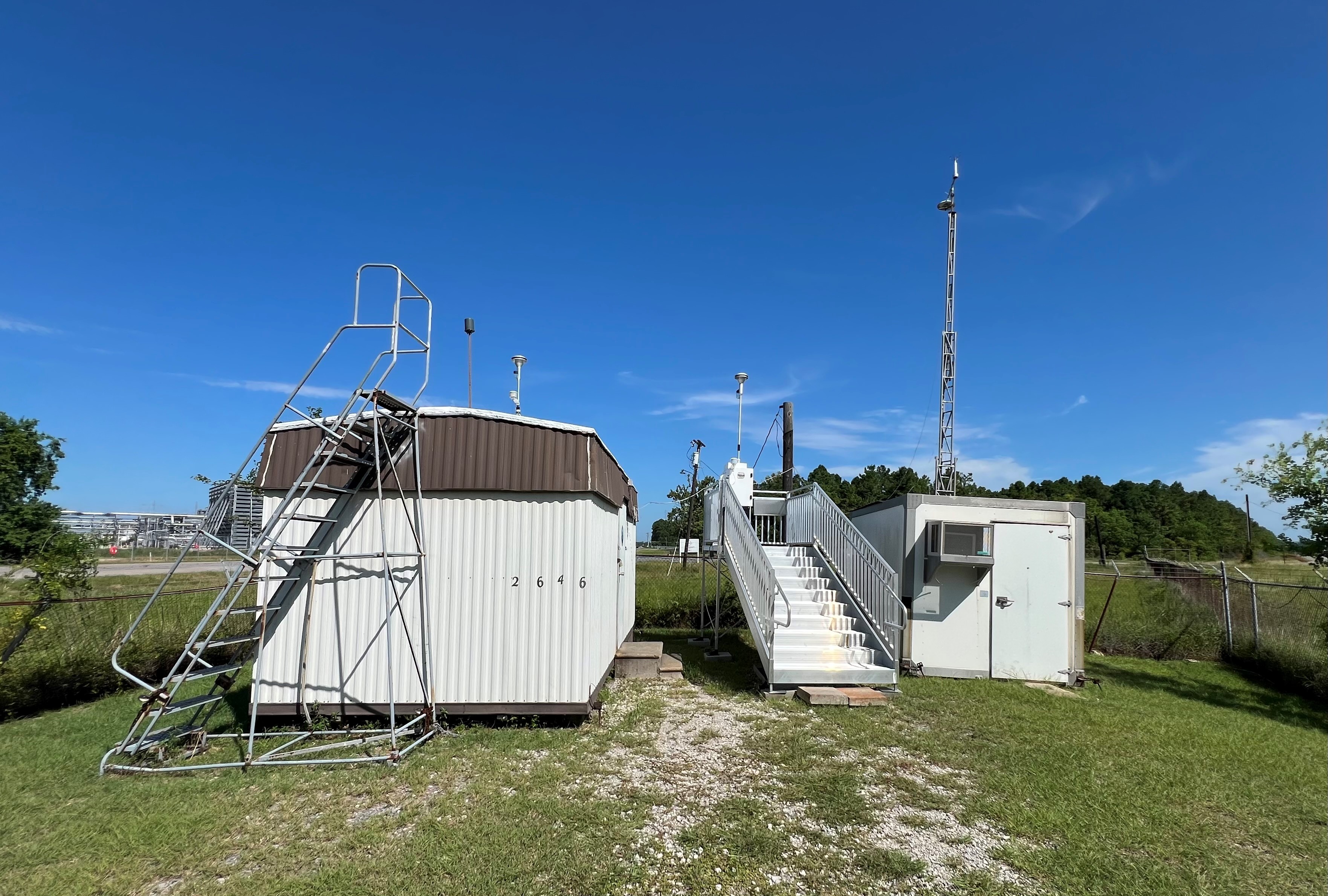 Two small metal buildings, one with a ladder to access the roof and the other with a metal platform with stairs holding a piece of air monitoring equipment with a blue sky in the background.