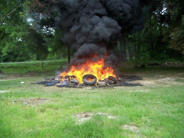 Picture of tires burning with dark black smoke on green grass with trees in back ground.